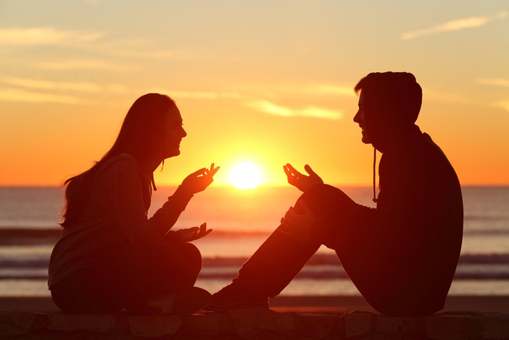 A man and a woman talking and laughing while sitting on the beach at sunset.