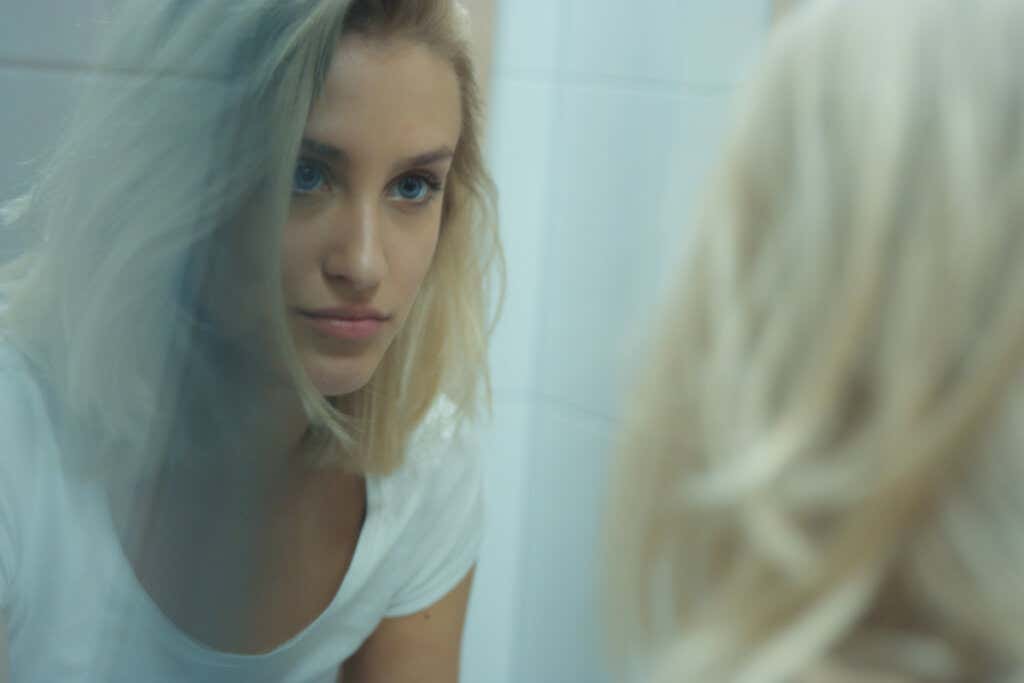 A woman with blond hair and blue eyes looking at herself seriously in the bathroom mirror.