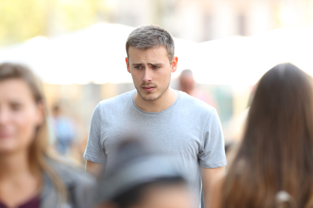Blond man in a gray shirt walks among several people and shows concern on his face