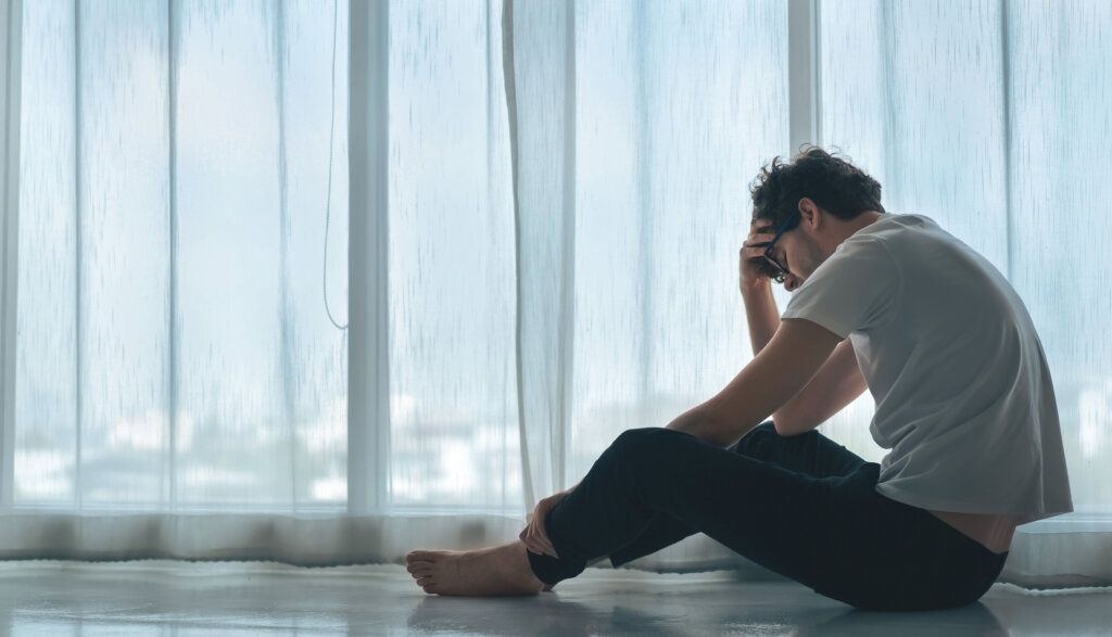 Homem preocupado sentado no chão ao lado de uma grande janela com cortinas brancas