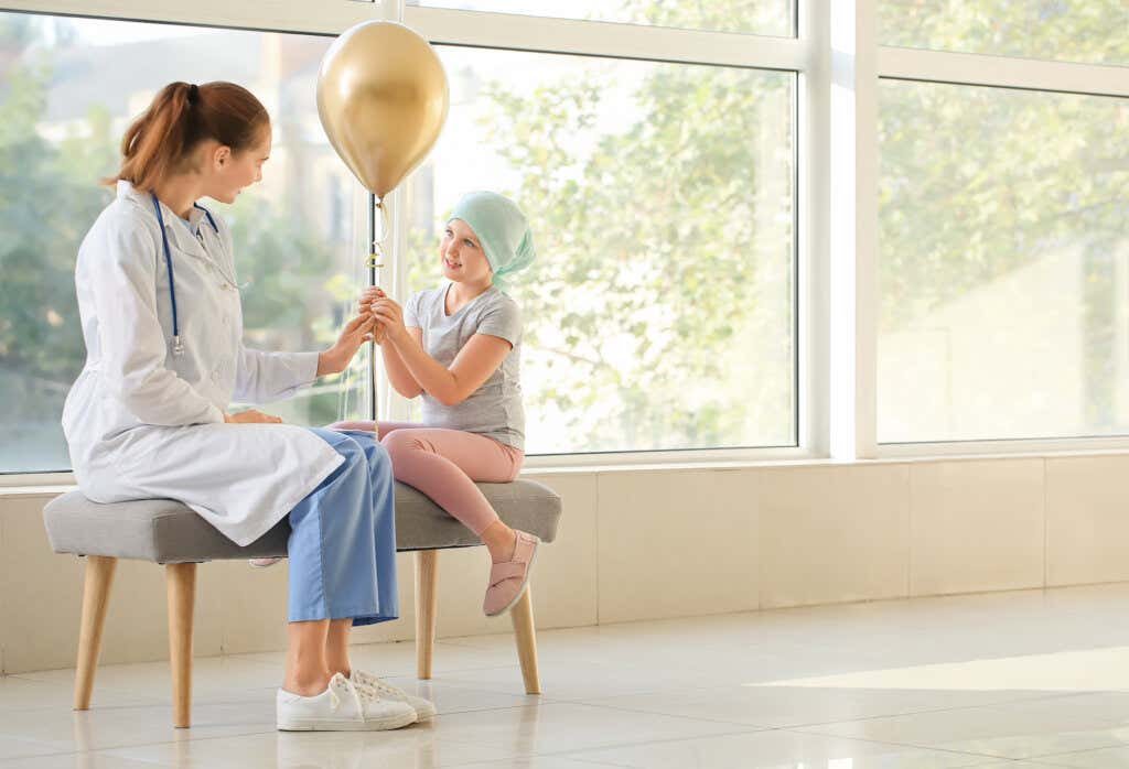 A girl with cancer holding a balloon and sitting on a bench in a hospital hallway, talking to a female doctor.