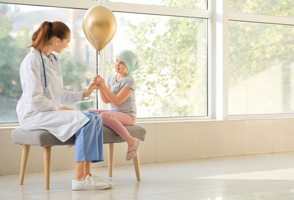 En flicka med cancer som håller en ballong och sitter på en bänk i en sjukhuskorridor och pratar med en kvinnlig läkare.