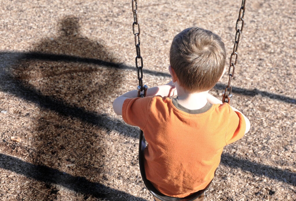 Boy sitting on his back on a swing while being watched by an adult who is reflected in the shadow