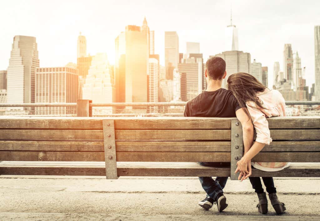 Couple holding hands sitting on a bench overlooking the city during sunset.