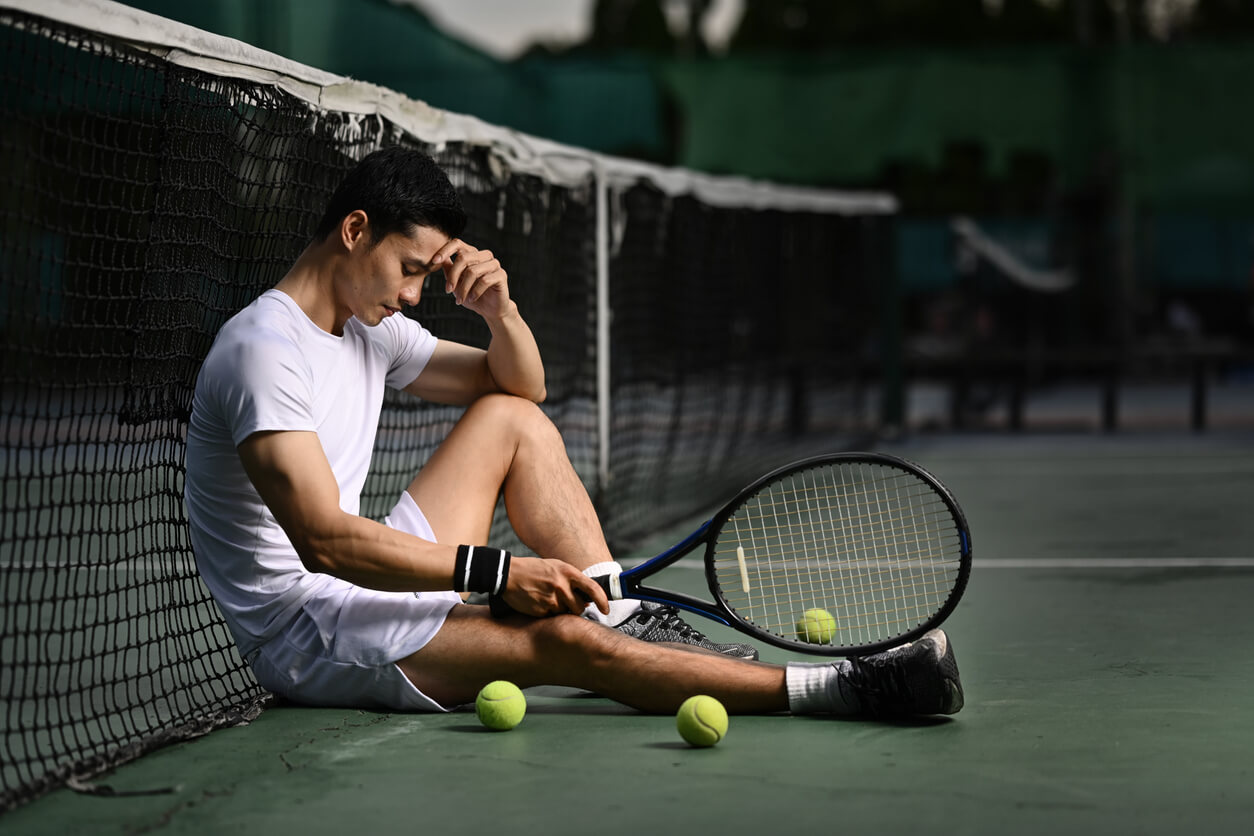 Jugador de tenis, sentado en la cancha, reflexiona sobre la pérdida de competencia. 