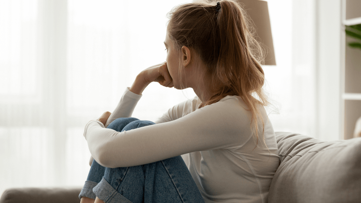 Mucha sentada en mueble mira hacia una ventana pensando en ideas negativas