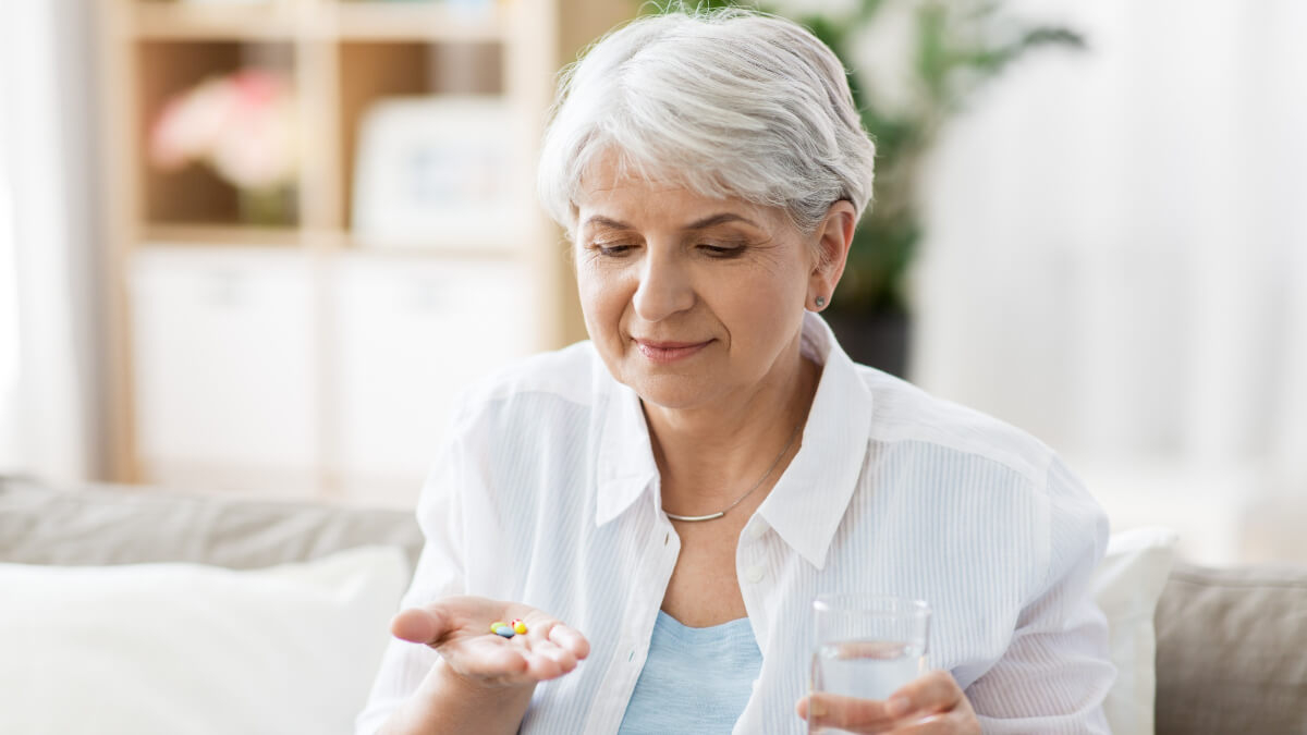 Mujer mayor sostiene suplementos en una mano y un vaso de agua en otra. 